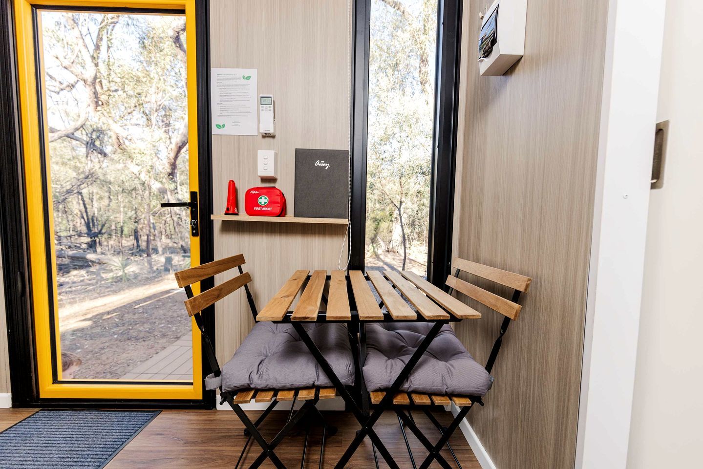 Une pièce avec une table et des chaises, une porte donnant sur l’extérieur et une petite étagère contenant une trousse de premiers secours et un objet rouge.