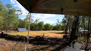 A covered outdoor area with a metal table and chairs, a solar panel, and a natural landscape with trees and a dirt path.