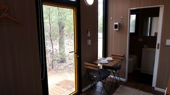 A room with a wooden table and chairs, a small kitchen area, and a door leading to an outdoor area with trees.
