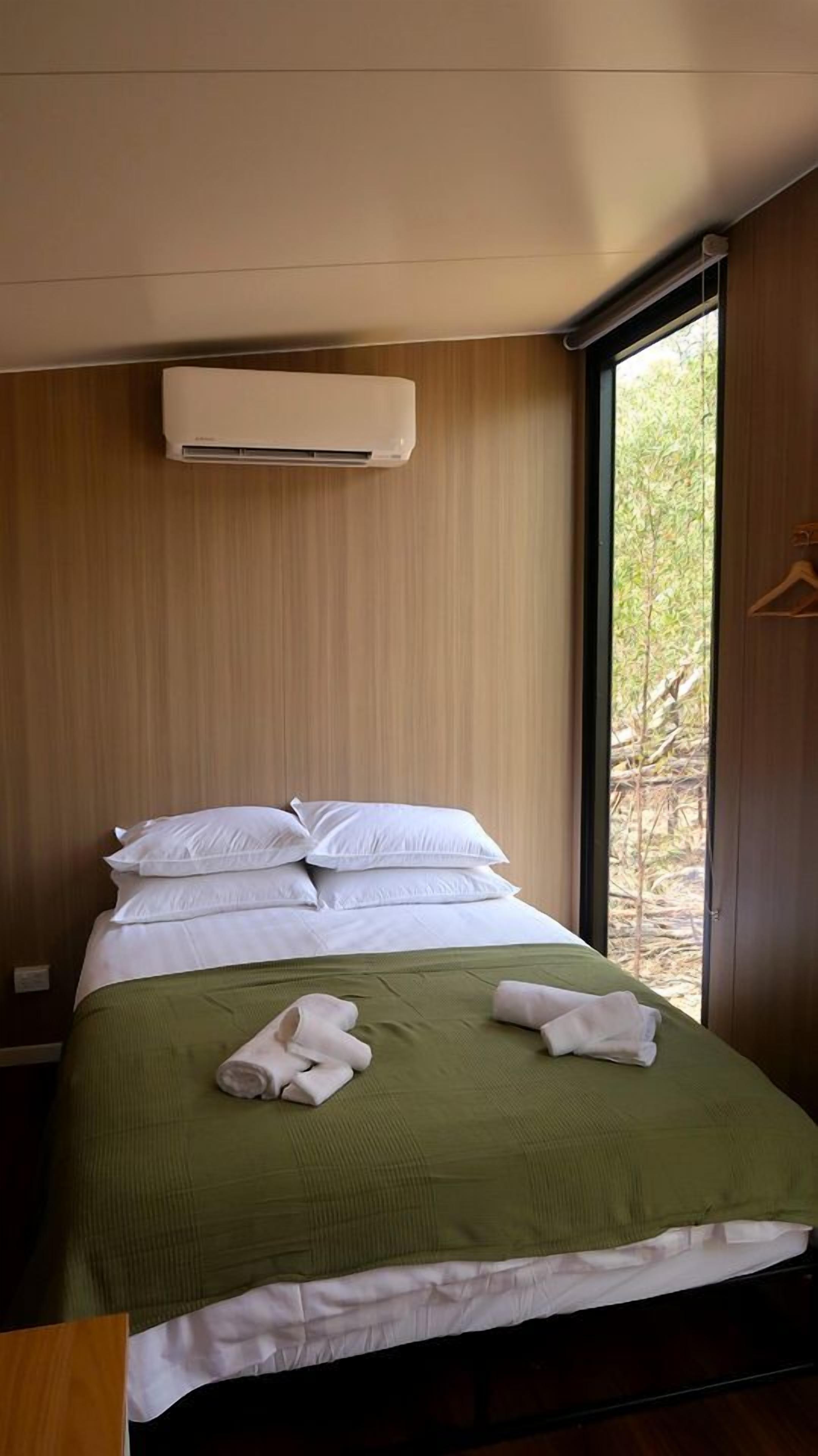 Ein Einzelbett mit einer grünen Tagesdecke und weißen Kissen, eine weiße Klimaanlage darüber und ein Blick auf Bäume durch ein Fenster.