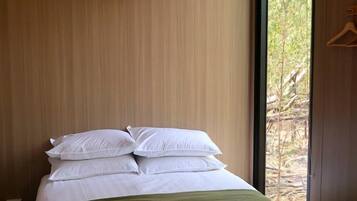 Ein Einzelbett mit einer grünen Tagesdecke und weißen Kissen, eine weiße Klimaanlage darüber und ein Blick auf Bäume durch ein Fenster.