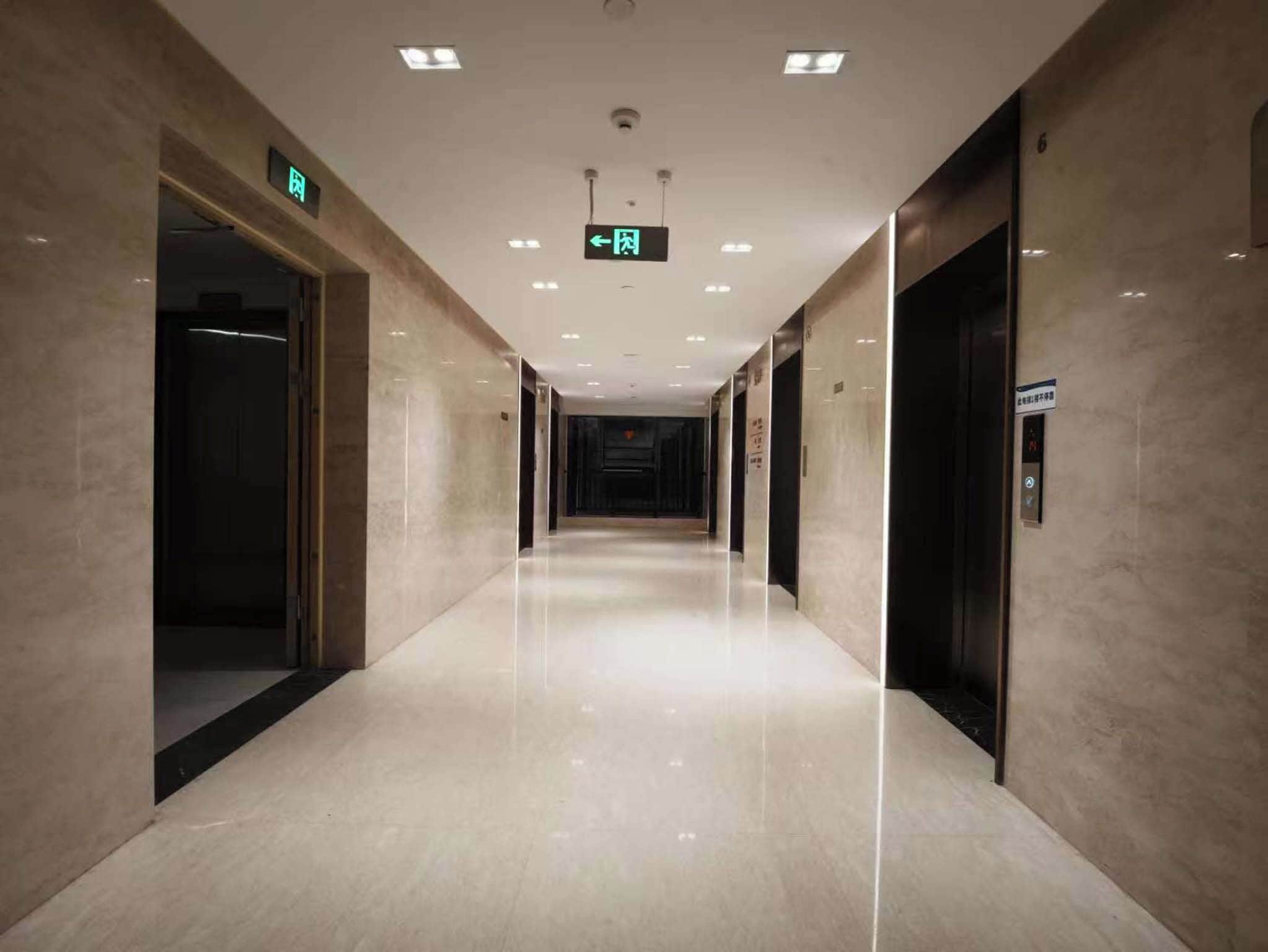 Un largo pasillo bien iluminado con piso pulido, puertas a ambos lados y un letrero de salida en el techo.