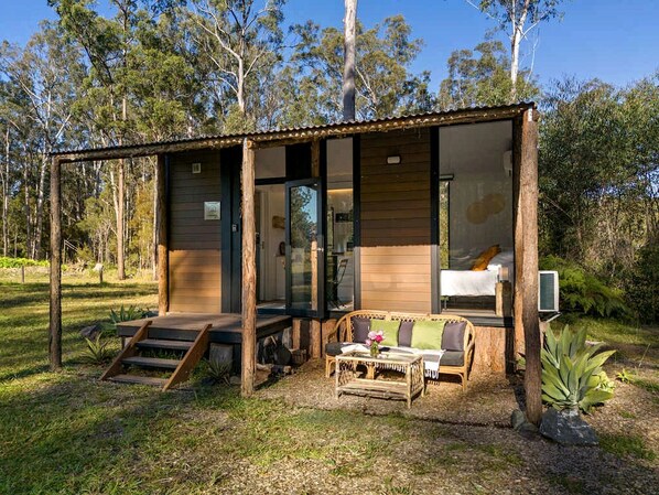 Una cabaña moderna con puerta de cristal, una cama y un pequeño rincón de descanso con mesa y sillas.