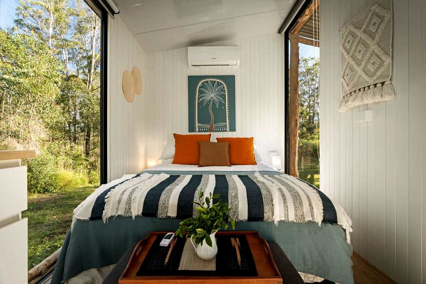 Un dormitorio con cama, almohadas, una mesita de noche con una planta y vistas al exterior.