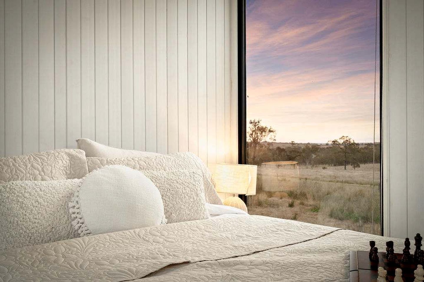 A bedroom with a large window, a bed with white bedding, and a lamp on the nightstand.