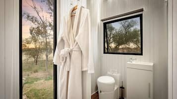 Un baño con ventana, inodoro blanco, lavamanos y una bata colgada en un perchero.