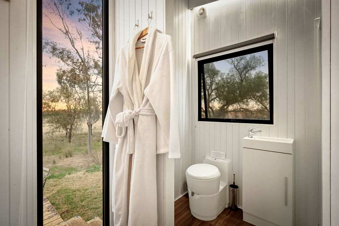 A bathroom with a window, a white toilet, a sink, and a robe hanging on a rack.