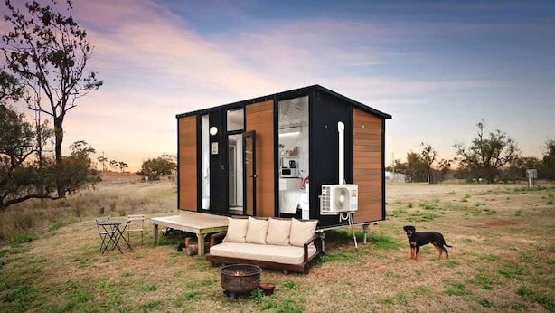 A modern, compact cabin with a sofa, a small table, and a fire pit, situated in a grassy field with trees in the background. - Charming Tiny House with Panoramic Views of the Valley in Queensland, Australia (Goombungee)