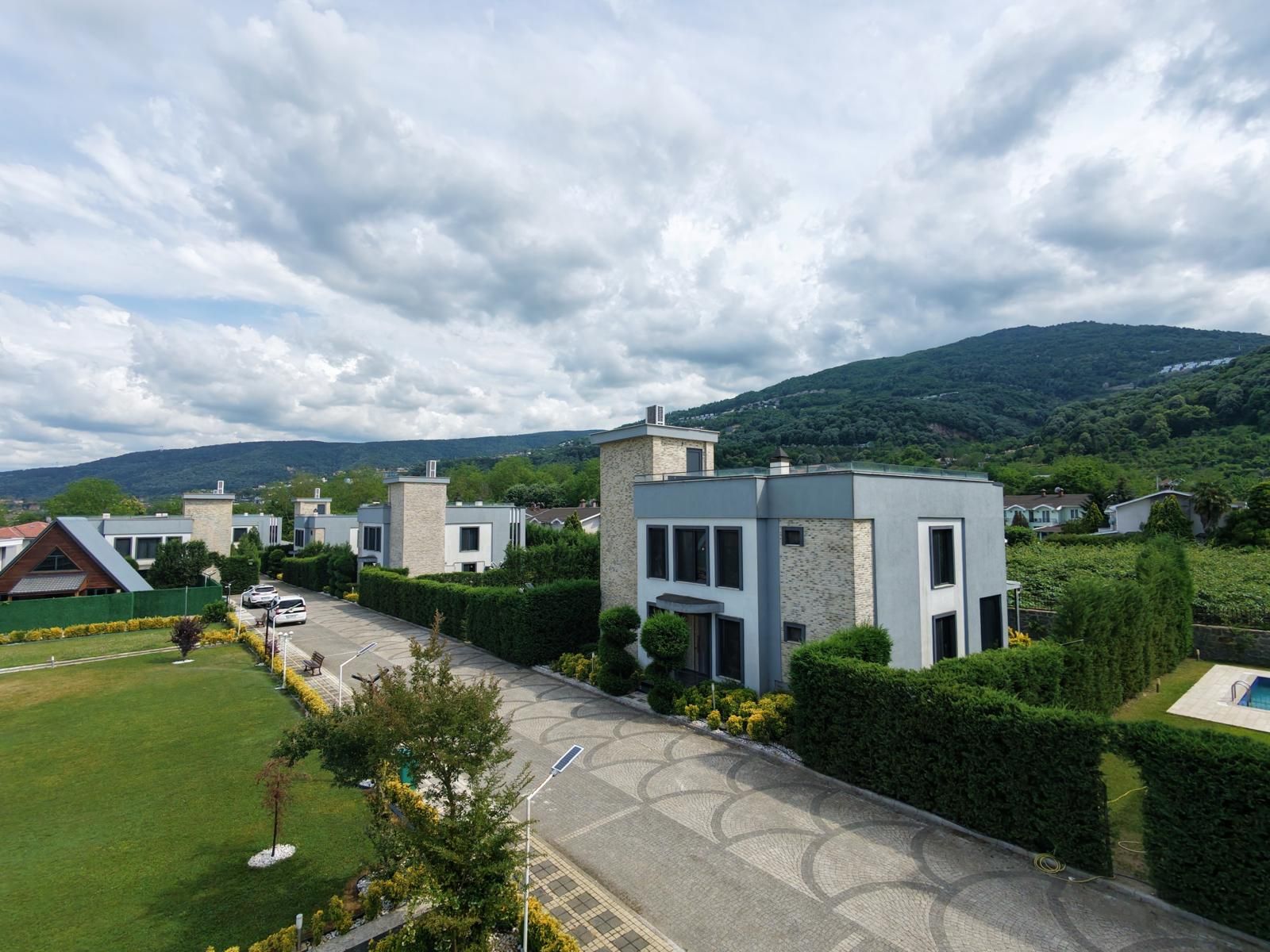 A residential area with modern houses, a paved driveway, and a hilly landscape in the background.