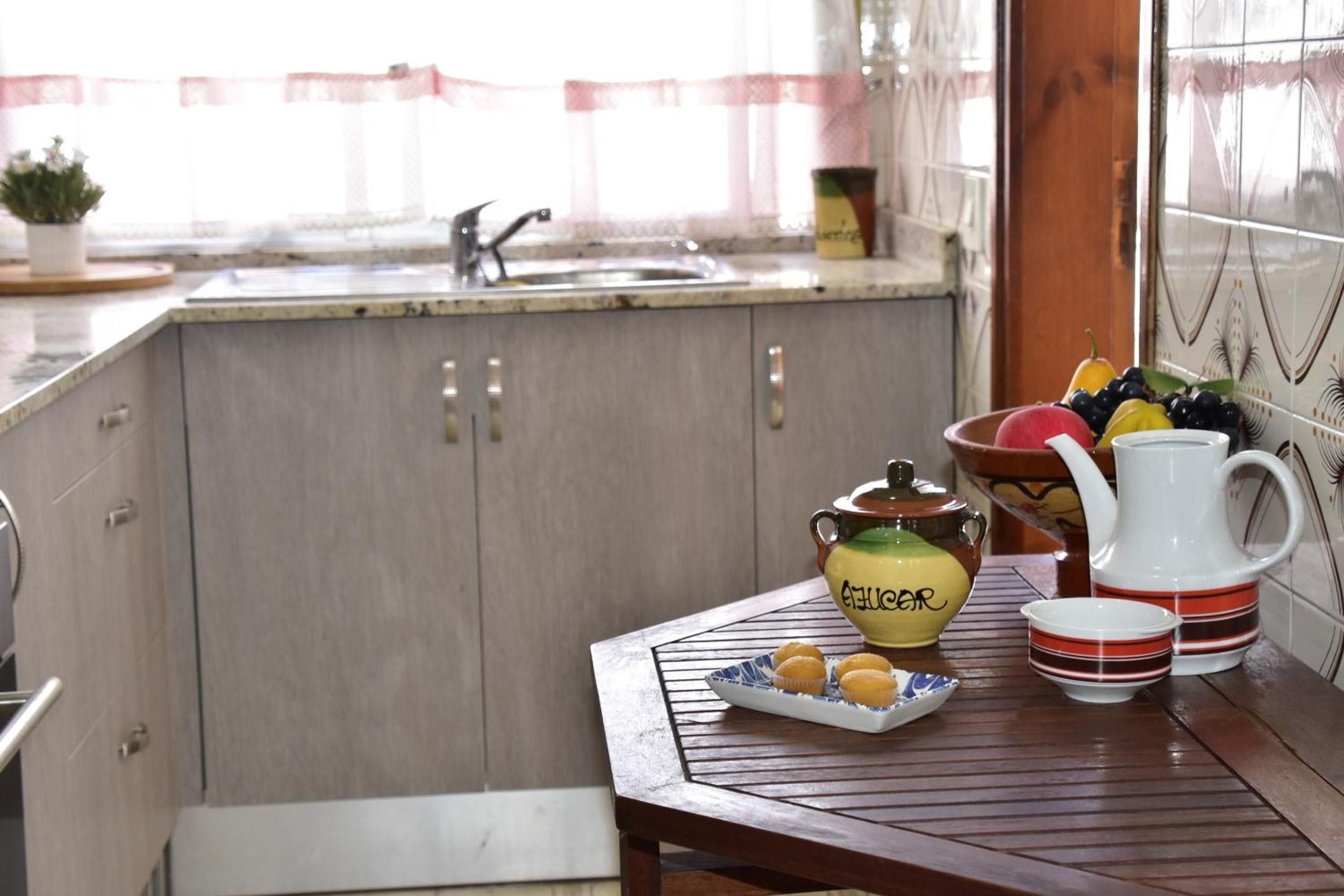 Una cocina con un lavamanos, armarios, un frutero, una tetera y una taza sobre una mesa.