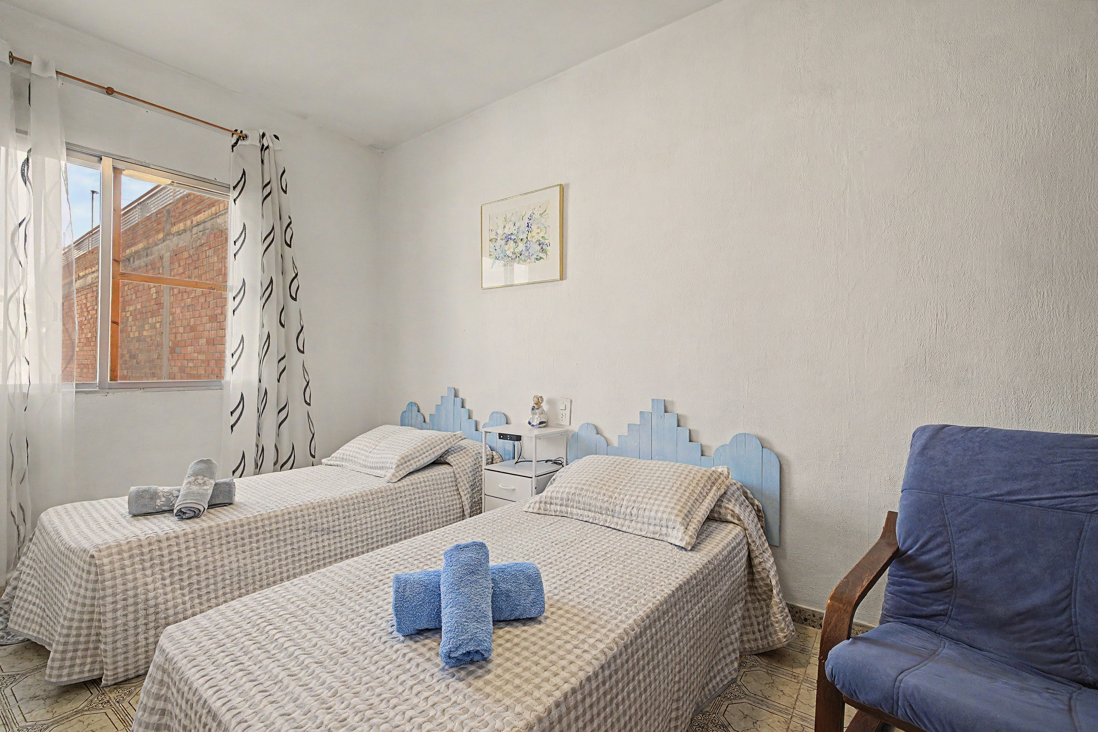 Quarto com duas camas, uma cadeira, escrivaninha e janela com cortinas.
