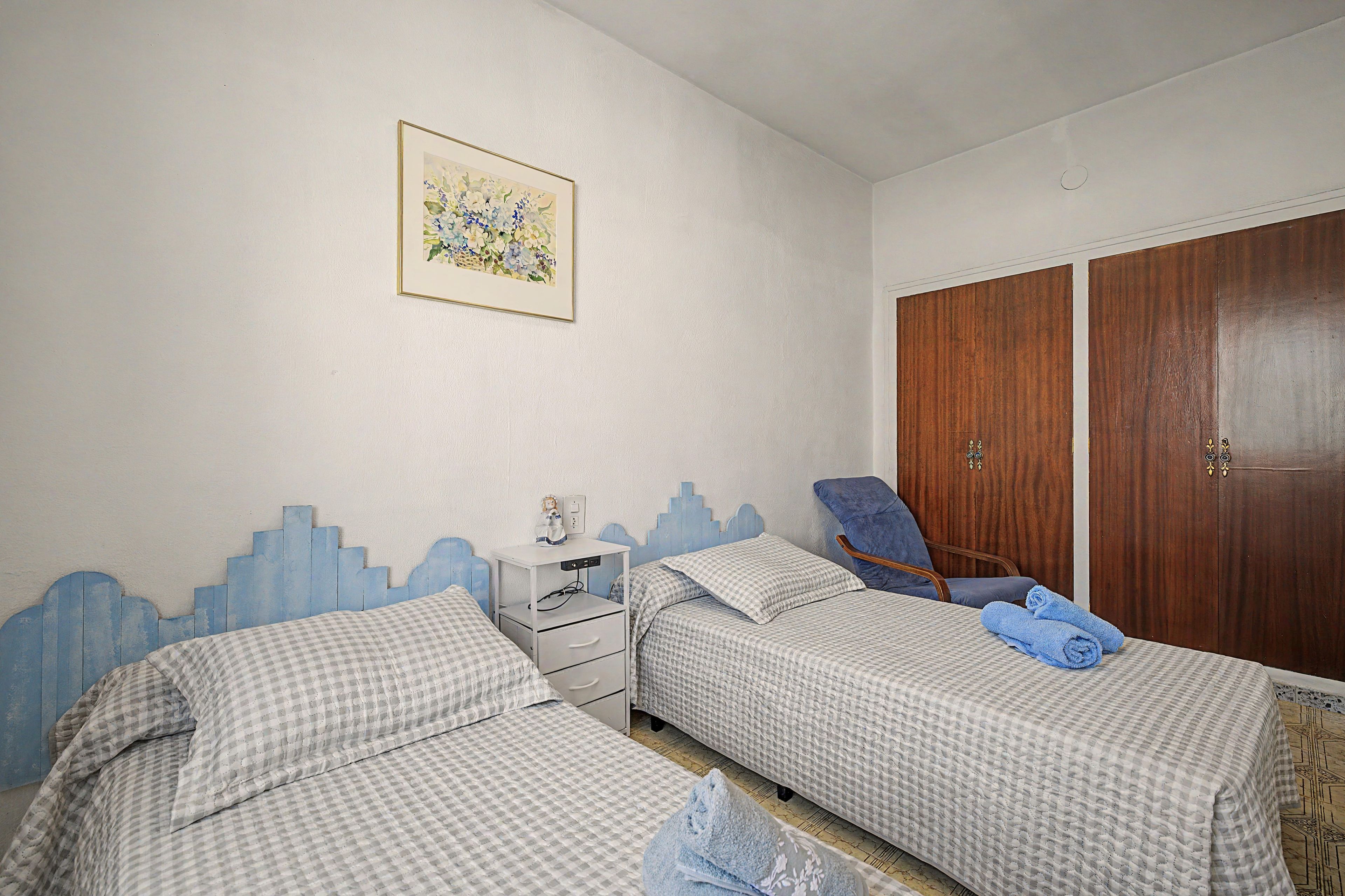 Un dormitorio con dos camas, una mesita de noche, una silla, un armario y un cuadro en la pared.