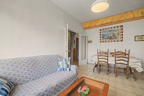 A bedroom with a bed, a table with a flower vase, a dining table with chairs, and a painting on the wall.