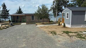 Ein Schotterweg führt zu einer Holzhütte mit grüner Tür, einem kleinen Gartenhaus und Blick auf einen See.