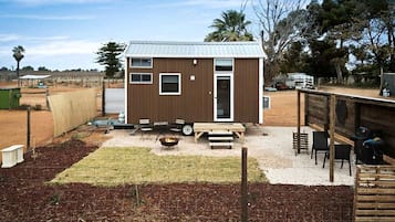 A small, single-story building with a flat roof, a door, and windows. There is a fire pit in the center, surrounded by a gravel area with a wooden deck and chairs.