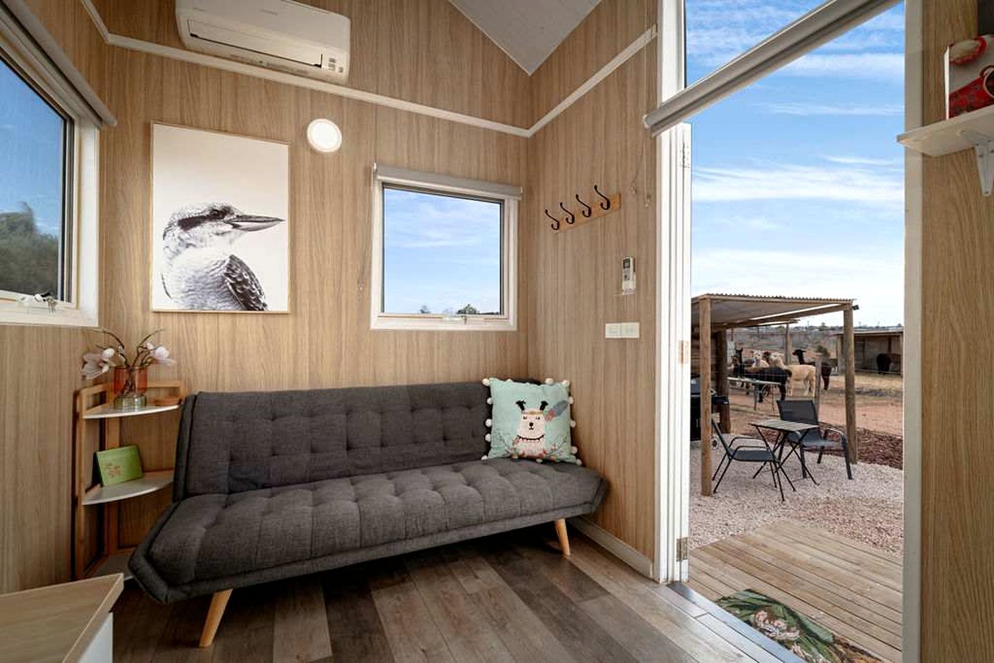 A room with a grey sofa, a wooden floor, a window, and a view of an outdoor area with animals.