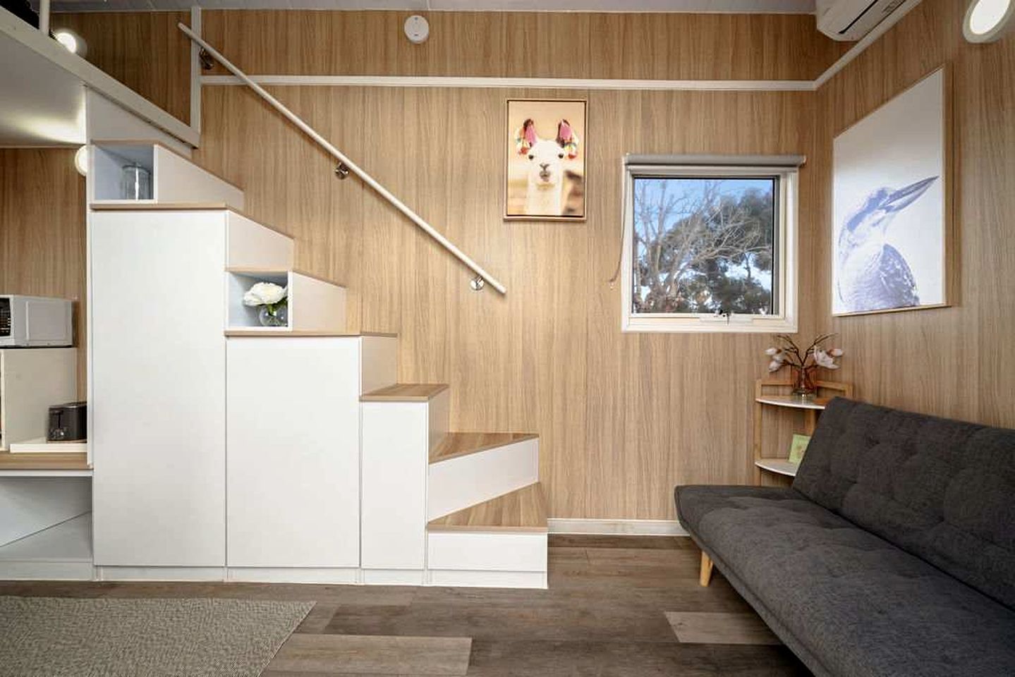 A modern interior with a staircase, a gray sofa, and a window with a view of trees.
