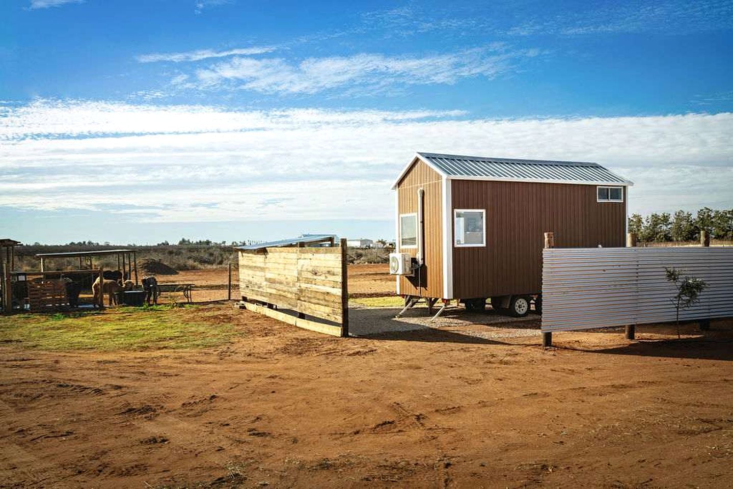 A small wooden structure with a metal roof, possibly a barn or storage shed, in a rural setting with a dirt ground and sparse vegetation.