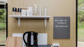 A room with a white shelf holding various glassware and mugs, a black kettle, a welcome sign, and a power outlet.