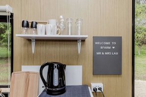 Une pièce avec une étagère blanche où sont disposés divers objets en verre et des tasses, une bouilloire noire, un panneau d’accueil et une prise électrique.