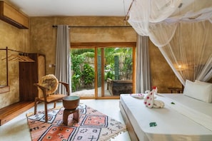 A room with a bed, a chair, a small table, and a view of a tropical garden through a sliding door. - Oke Ani Boutique Hotel (PAJE)