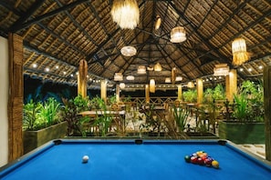 An indoor pool area with a thatched roof, surrounded by tables and chairs, and various hanging light fixtures. - Oke Ani Boutique Hotel (PAJE)