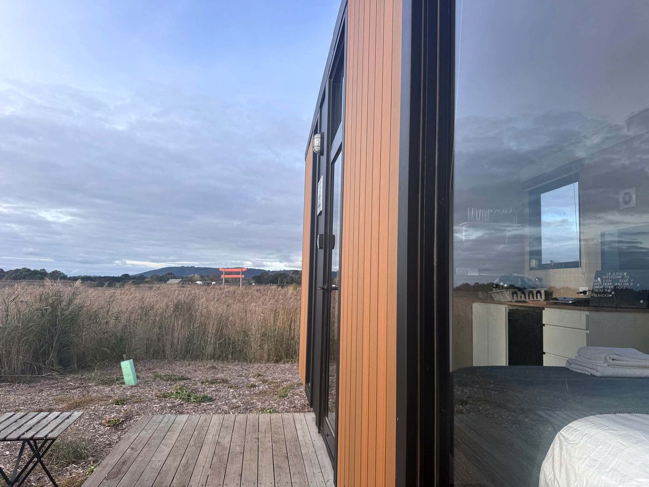 A modern building with large windows, a wooden deck, and a view of a field and distant hills.