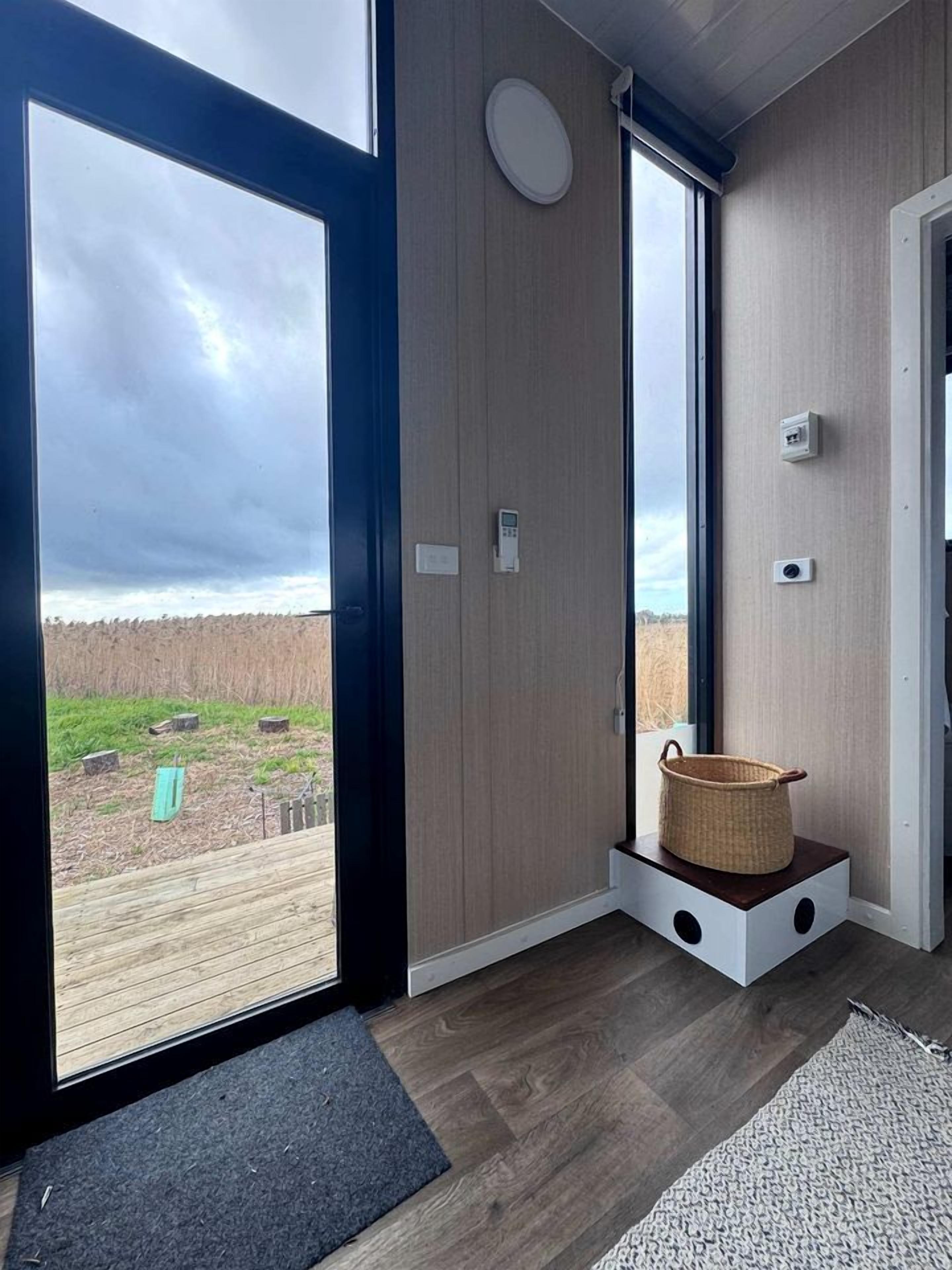 Une pièce dotée d’une grande porte-fenêtre coulissante donnant sur une terrasse, d’un petit banc en bois avec un panier, et d’un paillasson gris.