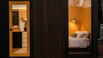 Una habitación compacta y moderna con una cama, lámpara de noche y una pequeña kitchenette visible a través de una puerta abierta.