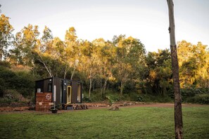 A small cabin with a wooden deck, surrounded by a grassy field and dense trees.