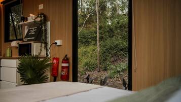A room with a bed, a small desk, a microwave, a fire extinguisher, and a window with a view of trees and foliage.