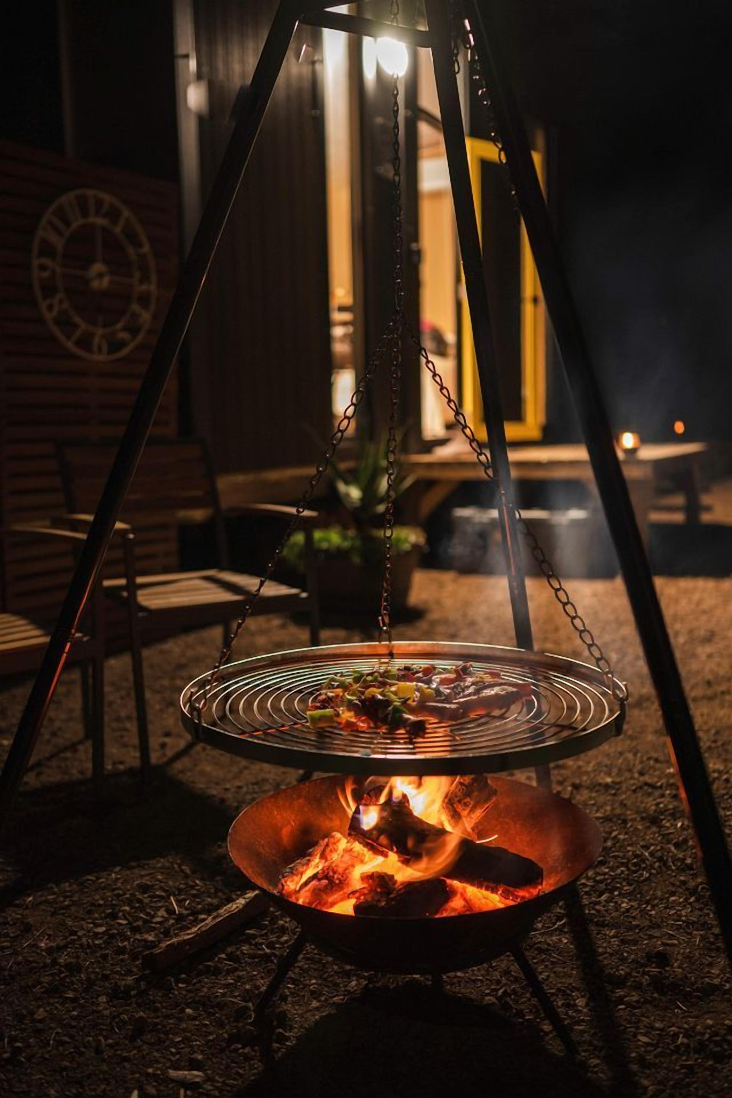 A portable grill with a fire burning underneath, set up outdoors with chairs and a clock in the background.