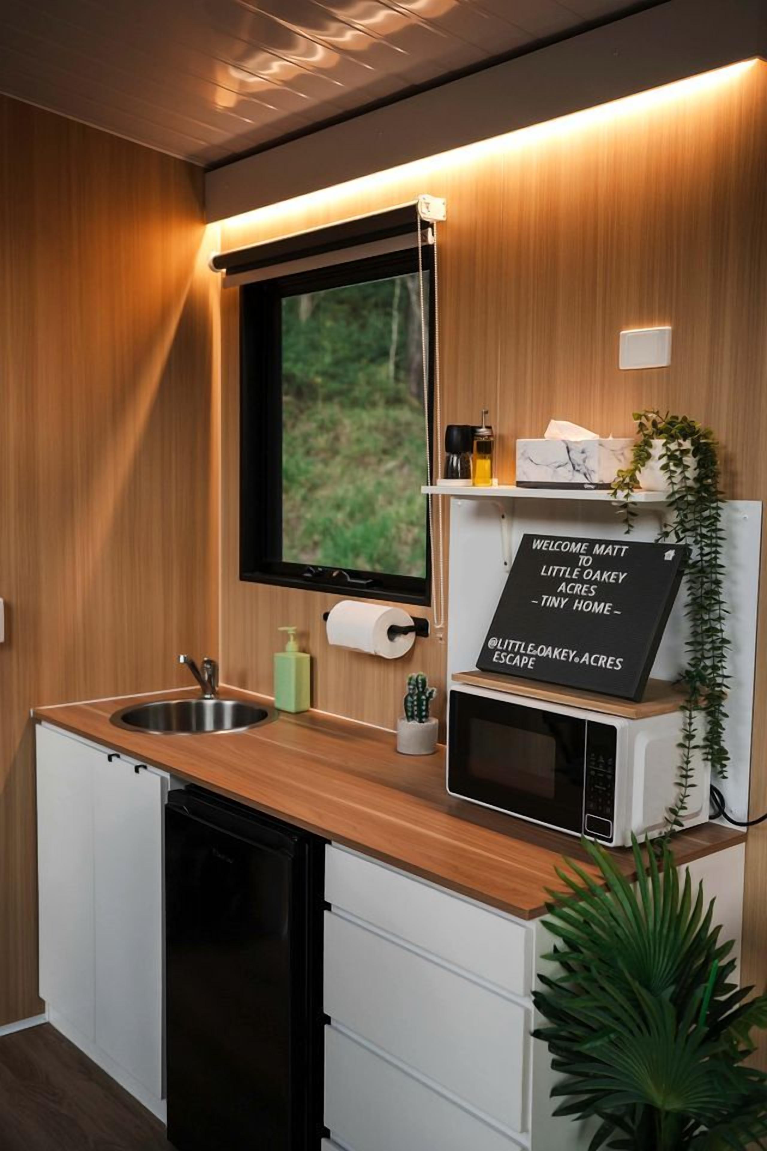 Een compacte keuken met een houten aanrecht, een spoelbak, een zwarte vaatwasser, een magnetron en een bordje dat Matt welkom heet in Little Oakey Acres Tiny Home Escape.