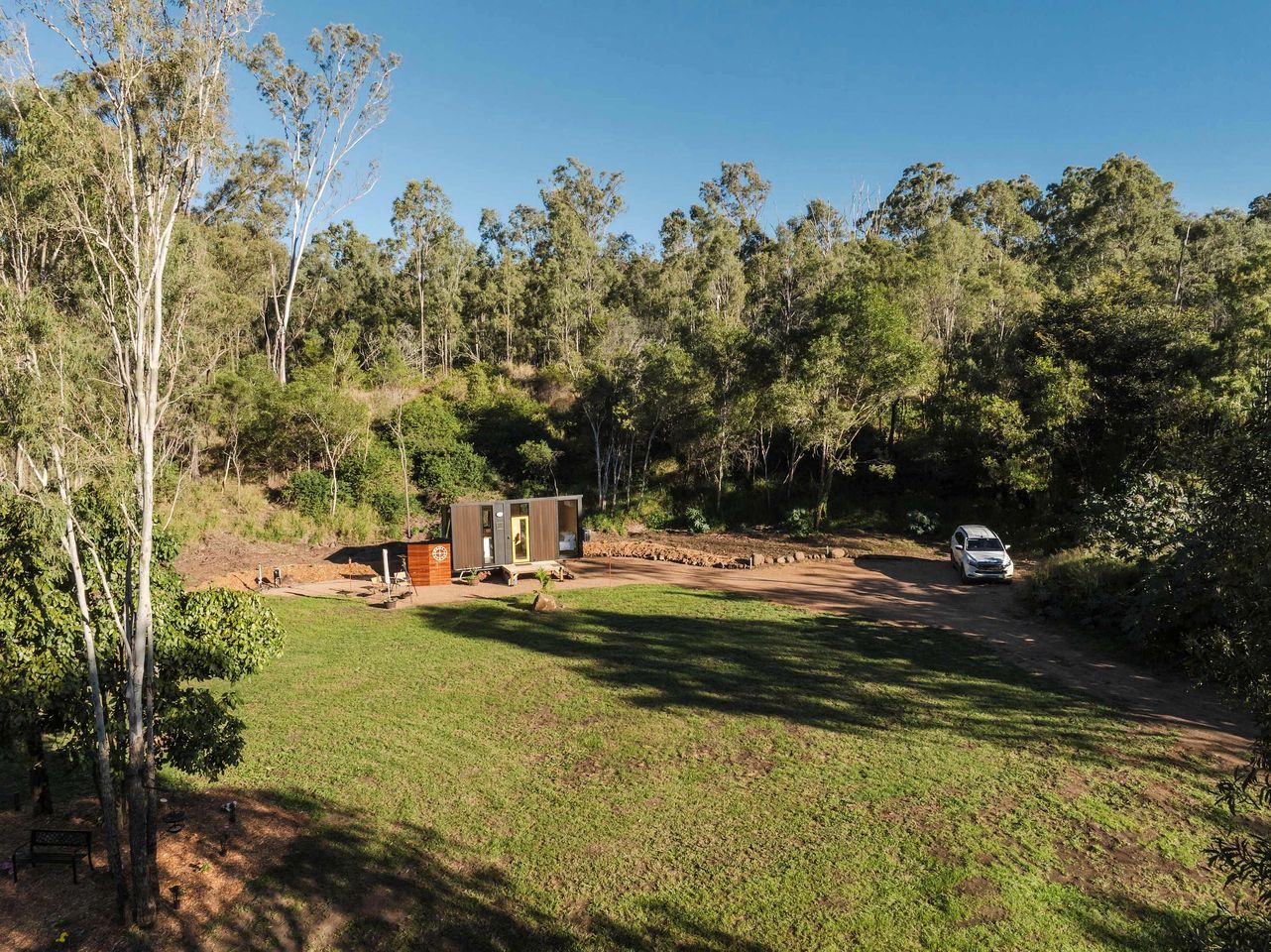 A campsite with a small wooden cabin, a paved area, and a dirt path leading to a vehicle.