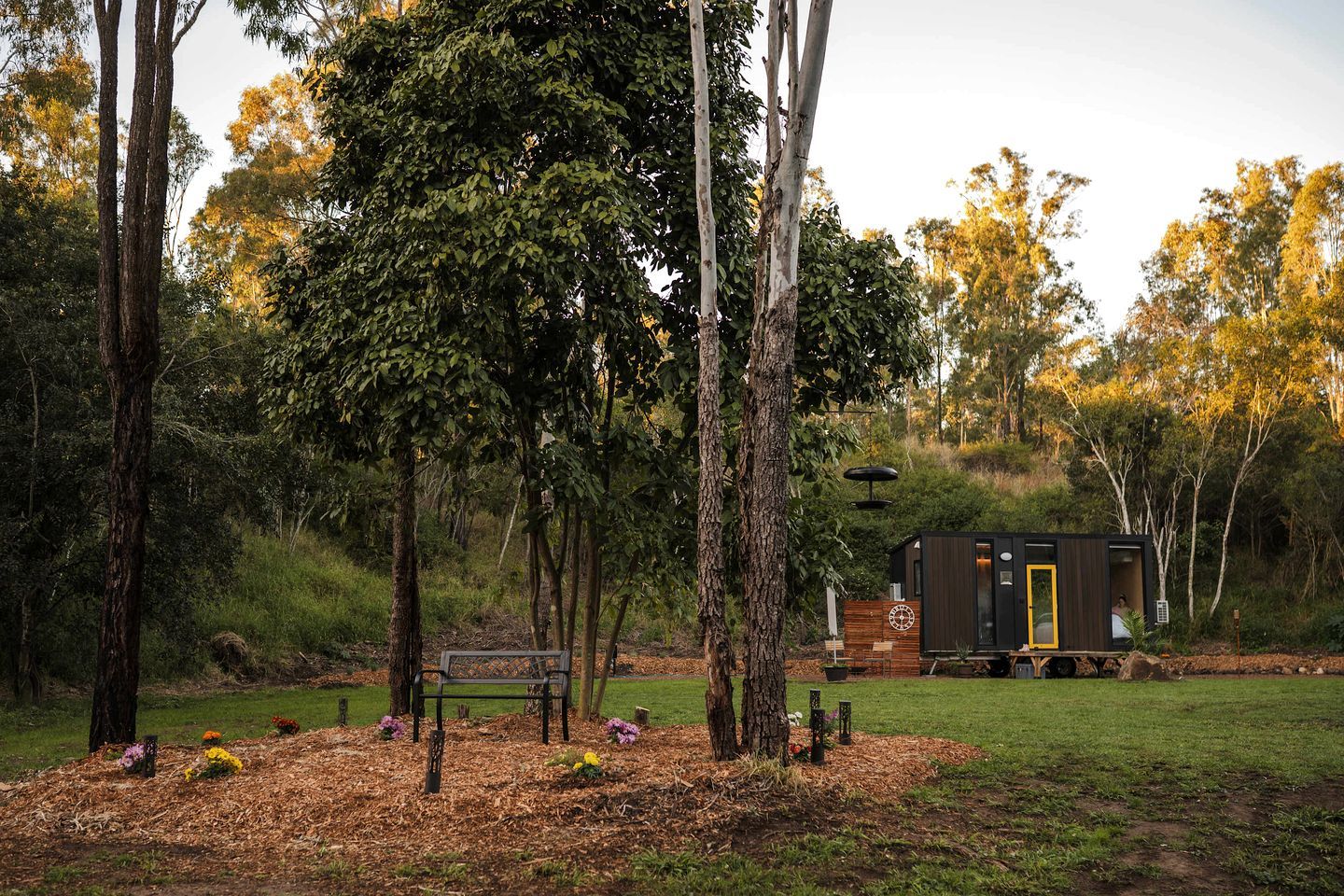 A cabin with a wooden deck and a bench, surrounded by trees and a grassy area.