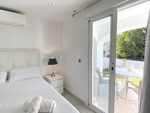Ein Schlafzimmer mit Bett, Nachttisch, Lampe und einer Glastür, die zu einem Außenbereich mit Tisch und Stühlen führt.