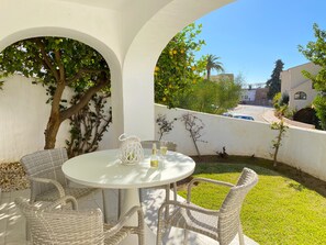 Ein runder Tisch mit Korbstühlen, einer weißen Karaffe und einer Champagnerflasche in einer sonnigen, grünen Umgebung mit einer weißen Wand.