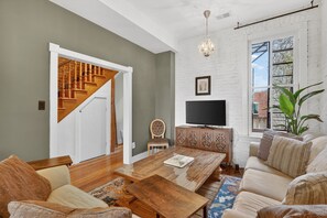 Un salon avec un escalier en bois, un téléviseur posé sur un meuble en bois et une grande fenêtre donnant sur l’extérieur.