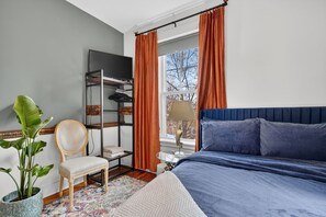 Une chambre à coucher avec un lit, une chaise, une étagère avec des vêtements, une plante et une fenêtre avec des rideaux.