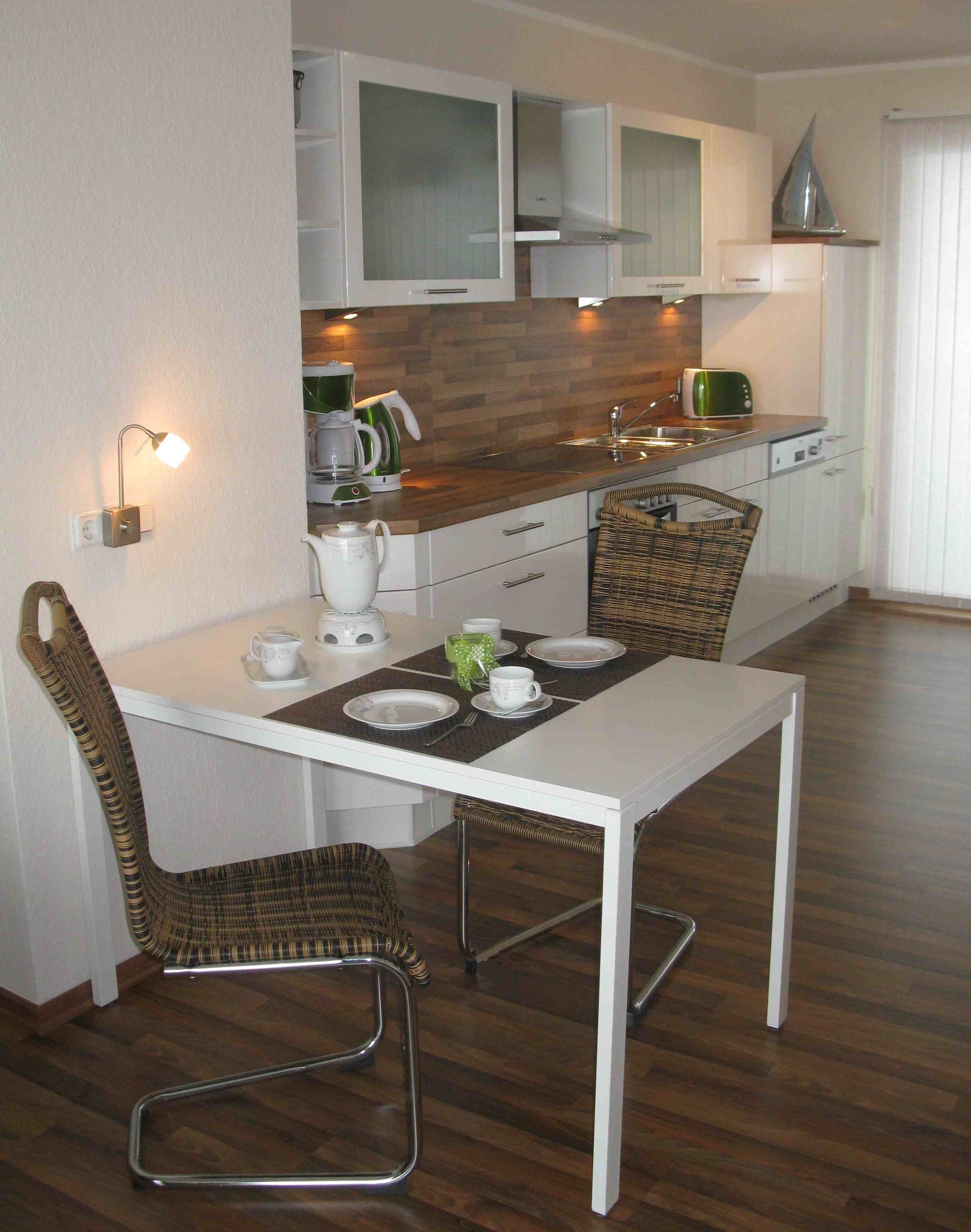 Une cuisine moderne avec des meubles blancs, une table à manger pour deux personnes et une chaise en osier.