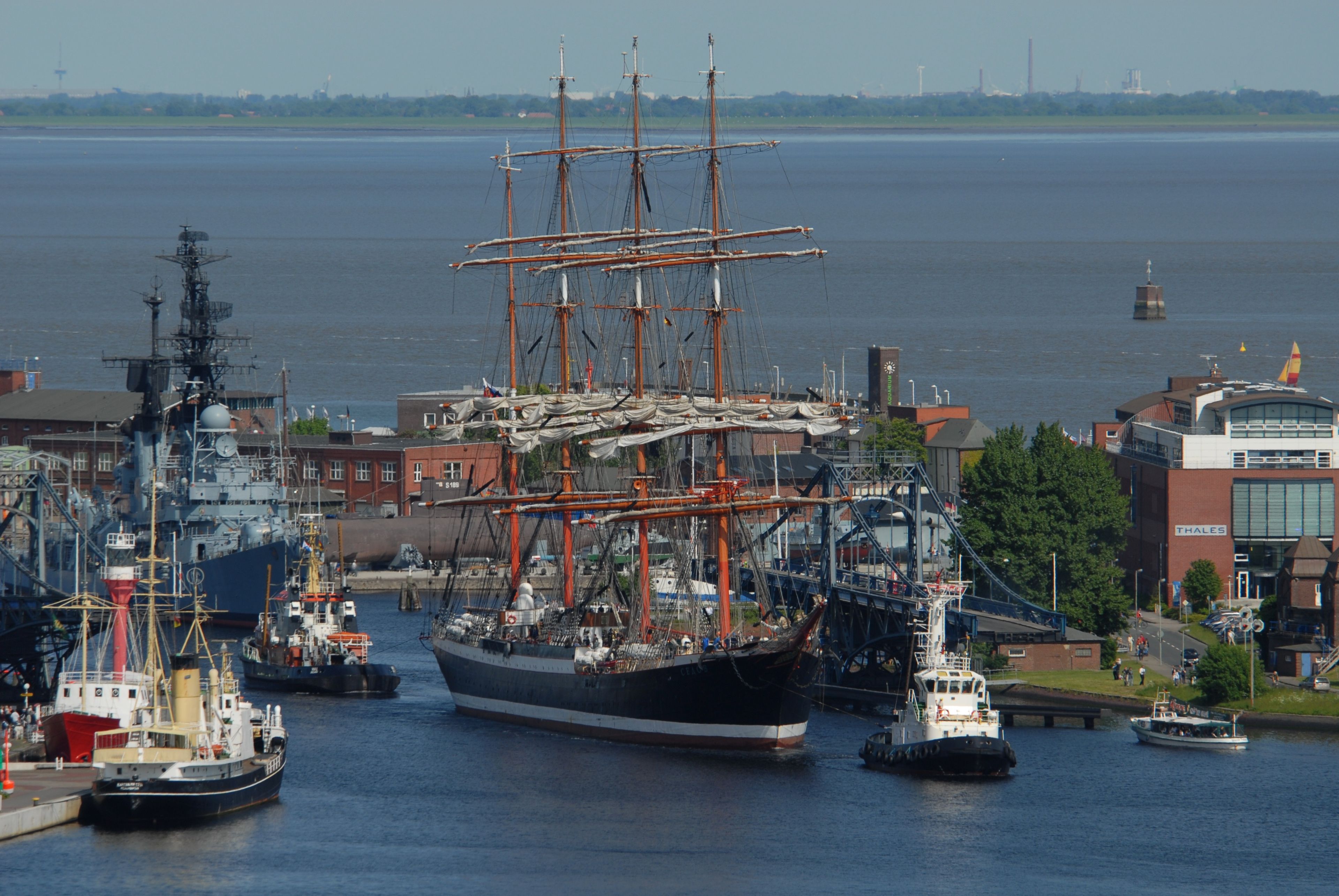 A large sailing ship docked at a port with industrial buildings and a bridge in the background.