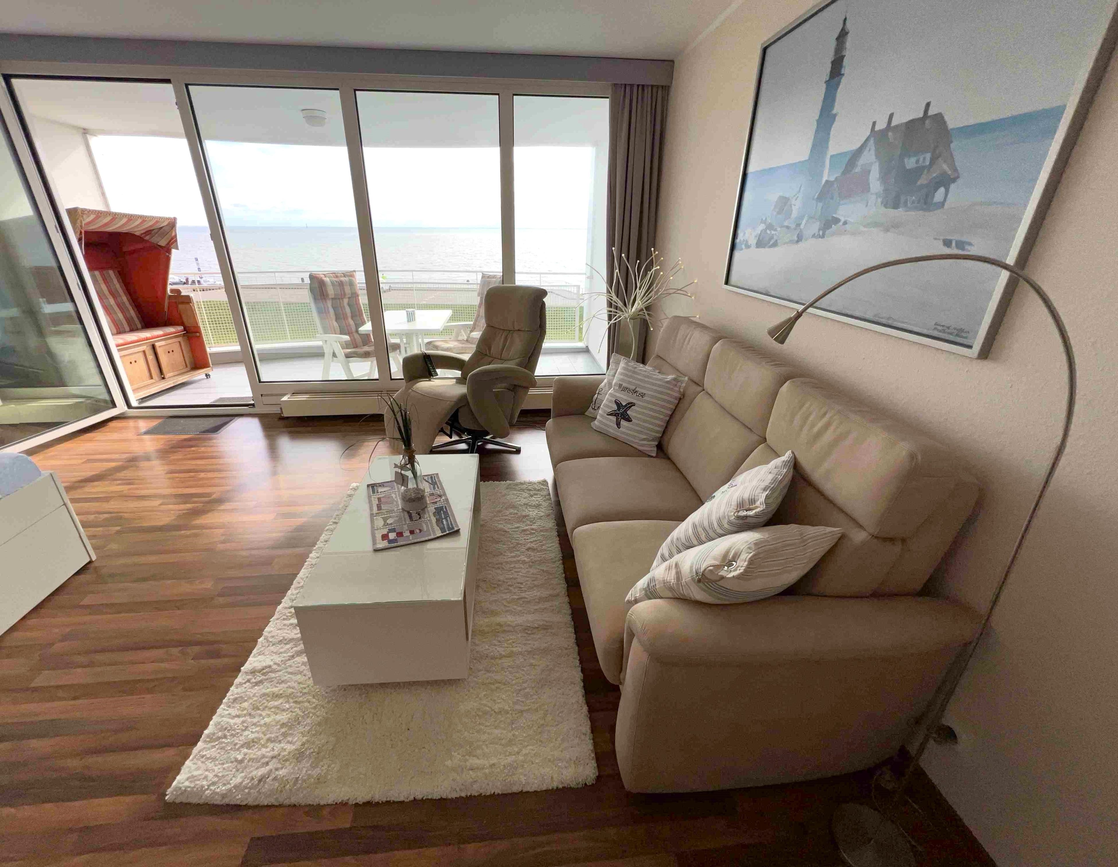 Ein modernes Wohnzimmer mit einer Couch, einem Couchtisch und einem Sessel. Ein großes Fenster bietet Blick auf das Meer.