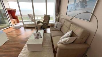Ein modernes Wohnzimmer mit einer Couch, einem Couchtisch und einem Sessel. Ein großes Fenster bietet Blick auf das Meer.