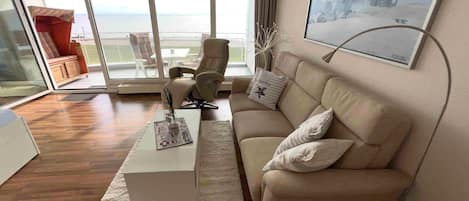 A modern living room with a sofa, a coffee table, and a chair. There is a large window with a view of the sea.