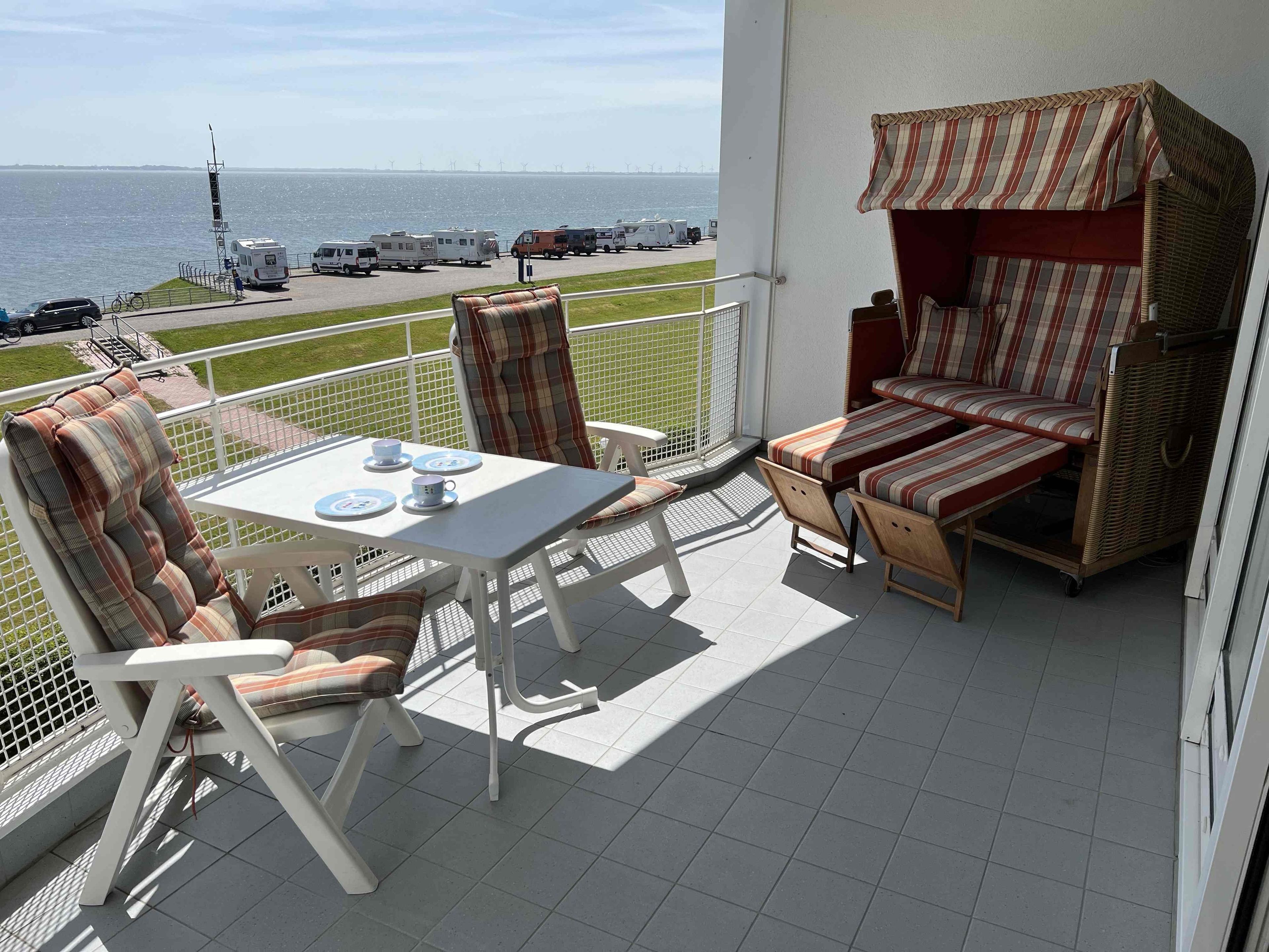 Ein Balkon mit Tisch und Stühlen, einem Korbsessel und Blick auf das Meer und Windkraftanlagen.
