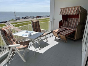 Ein Balkon mit Tisch und Stühlen, einem Korbsessel und Blick auf das Meer und Windkraftanlagen.