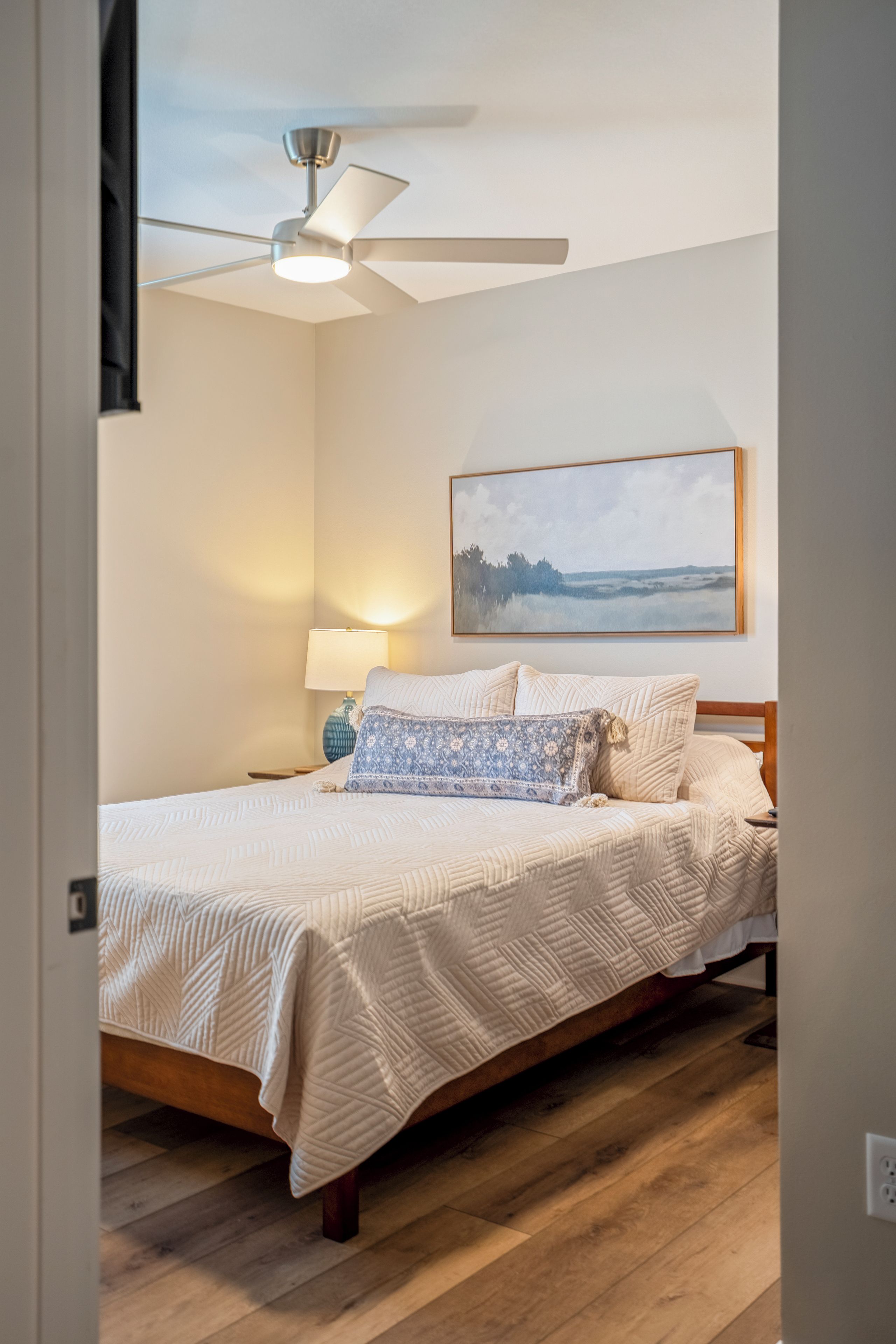 A bedroom with a bed, a ceiling fan, a lamp, and a painting on the wall.