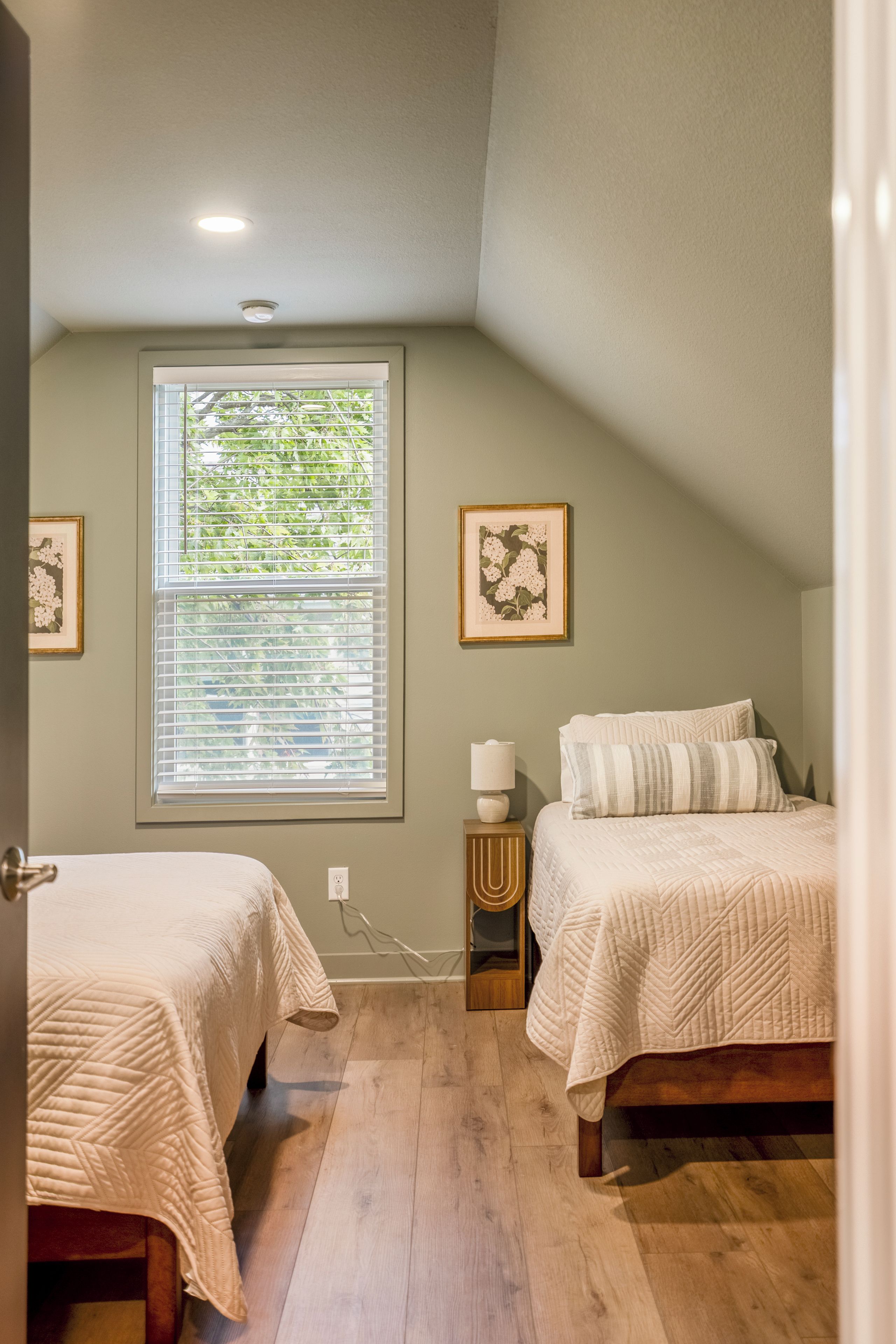 A bedroom with two beds, a window with blinds, a bedside table with a lamp, and framed artwork on the walls.