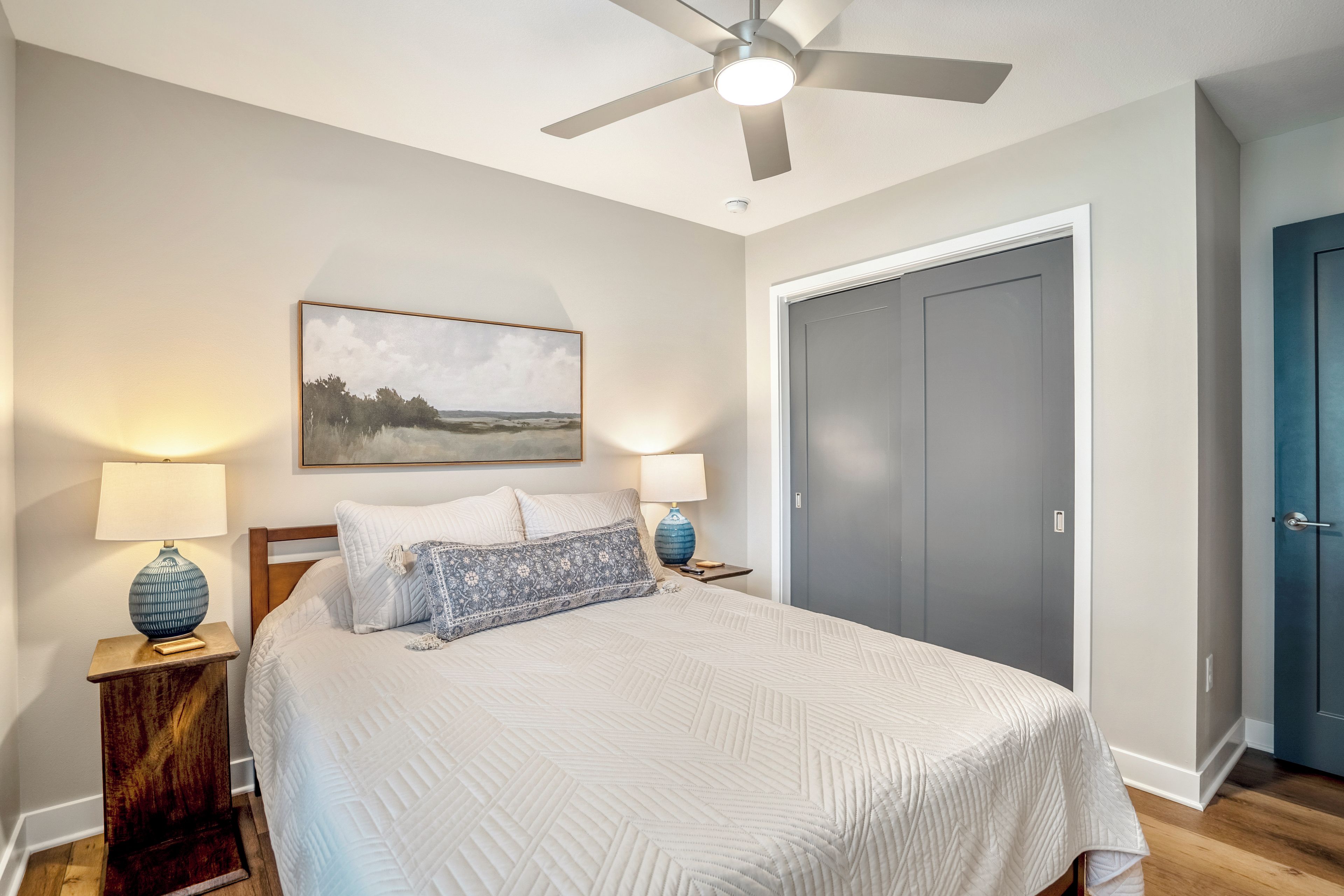 A bedroom with a bed, two bedside lamps, a ceiling fan, a framed landscape painting, and a wardrobe.