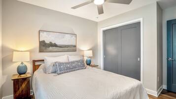 A bedroom with a bed, two bedside lamps, a ceiling fan, a framed landscape painting, and a wardrobe.